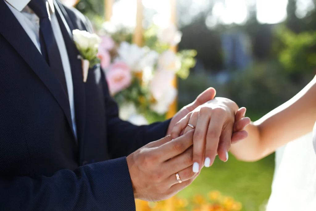 Close-up of bride and groom exchanging wedding rings outdoors, symbolizing second marriage commitment, with floral background.